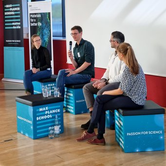 The panel discussion reminded us to make a career choice that we ourselves are happy with and can relate to Four individuals seated on blue cubes during a panel discussion, surrounded by banners from Max Planck School Matter to Life and DWI Institute.