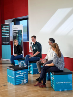 Four individuals seated on blue cubes during a panel discussion, surrounded by banners from Max Planck School Matter to Life and DWI Institute.