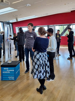 Students gathered in a contemporary setting with red walls and large windows, interacting around display panels with educational content. A small seating cube with "Max Planck School Matter to Life" branding is in the foreground.