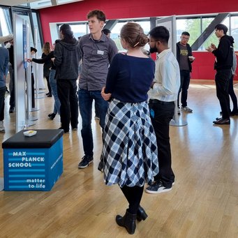 Students gathered in a contemporary setting with red walls and large windows, interacting around display panels with educational content. A small seating cube with "Max Planck School Matter to Life" branding is in the foreground.