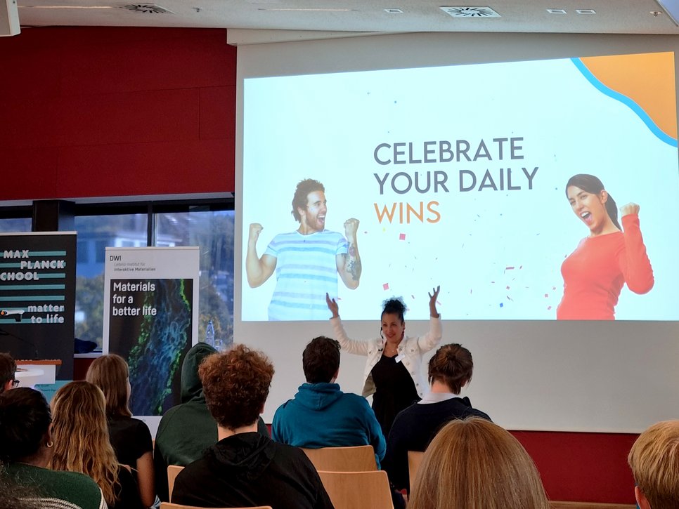 A seminar room with attendees focused on a presentation slide encouraging the celebration of daily wins. The room features banners for Max Planck School  Matter to Life and DWI, Leibniz Institute for Interactive Materials.