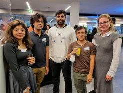 Five people dressed casually with visible name tags, standing together in a modern indoor setting. Two hold drinks, suggesting a social gathering or networking event.