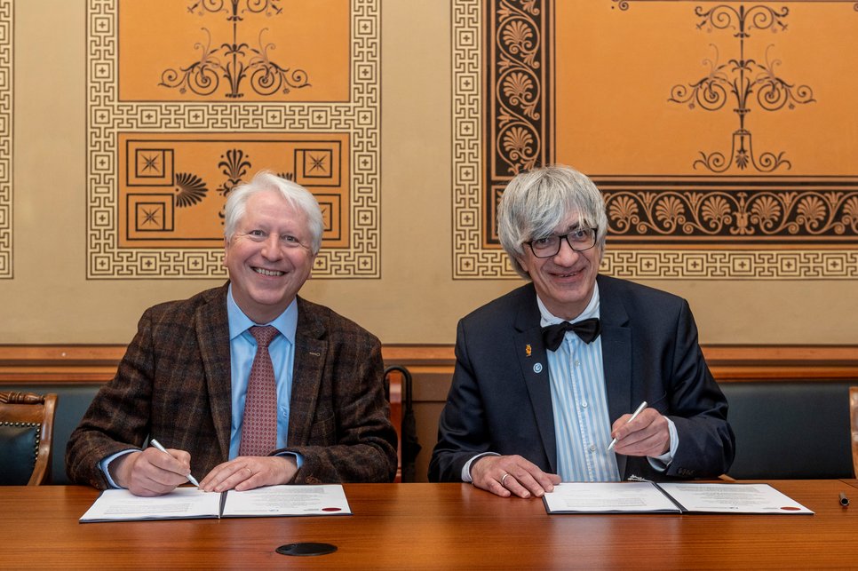 Prof. Metin Tolan and Prof. Bernhard Eitel are sitting at a desk, ready to sign the contract, and are smiling into the camera.
