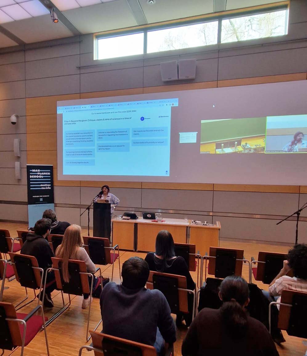 A person is giving a presentation to an audience in a modern conference room. The screen shows presentation slides and a video call with virtual participants. A banner is standing to the left of the lectern.