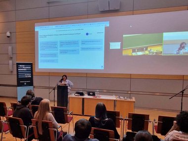 A person is giving a presentation to an audience in a modern conference room. The screen shows presentation slides and a video call with virtual participants. A banner is standing to the left of the lectern.