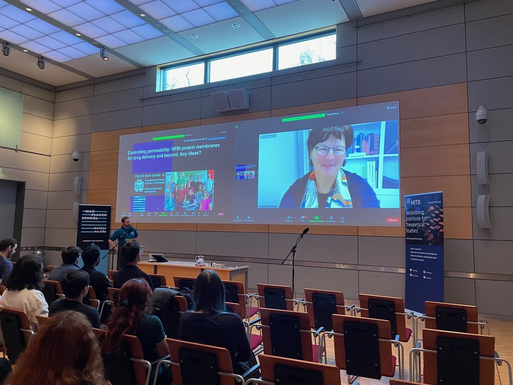 Attendees in a modern conference room observe a presentation on HFBI protein membranes for drug delivery, with a large screen showing speaker slides and video call setup.