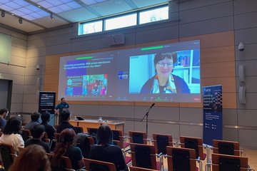 Attendees in a modern conference room observe a presentation on HFBI protein membranes for drug delivery, with a large screen showing speaker slides and video call setup.