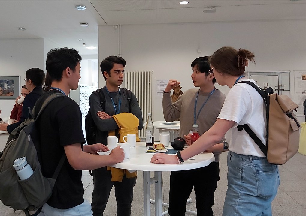 A group of students is standing around a table, drinking coffee and discussing