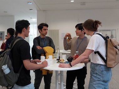 A group of students is standing around a table, drinking coffee and discussing