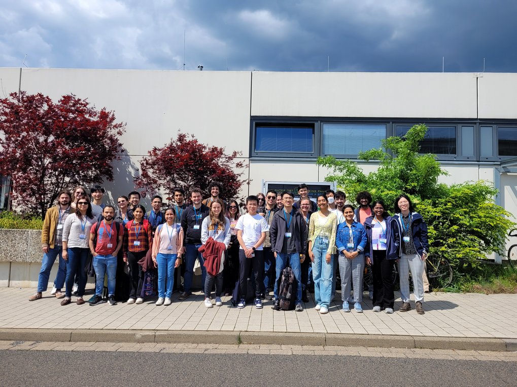 A large group of young individuals, dressed casually, gather on a paved area in front of a modern building, with red-leaved trees and green bushes in the background.