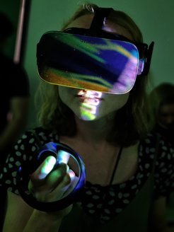 Close-up of a female student wearing VR goggles and holding one of the controllers raised in front of her. Different colors like orange, yellow or even purple illuminate the image.
