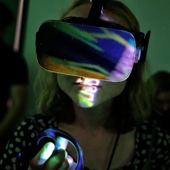 Is VR the future? In that case our students will not be left behind Close-up of a female student wearing VR goggles and holding one of the controllers raised in front of her. Different colors like orange, yellow or even purple illuminate the image.