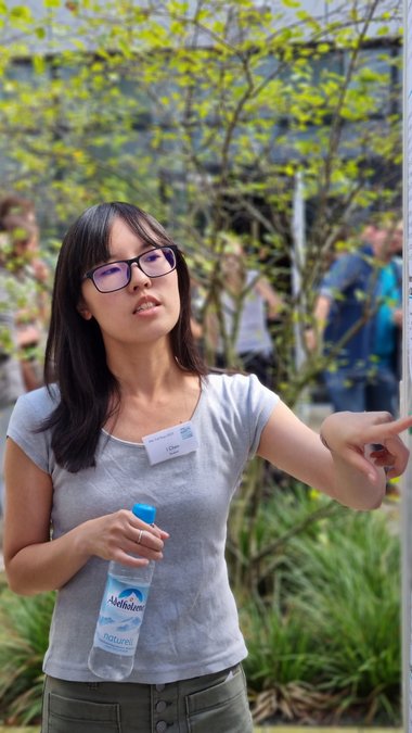 I Chen points to a spot on her poster with her left index finger and explains something to another student to her left who is looking at that spot. In her right hand she holds a water bottle. In the background, other people can be seen as well as greenery.