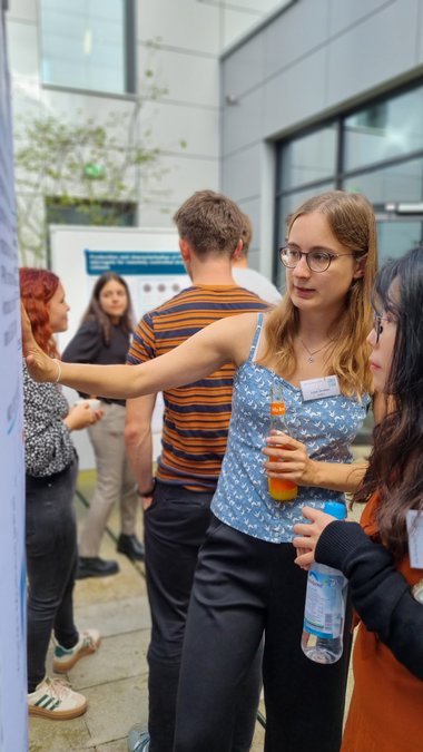 Lena Strieker stands with another student at her poster and explains the contents to her. Both are holding a small bottle in their hands. In the background, other participants and also another poster can be seen, as well as the walls of the building in whose courtyard they are.