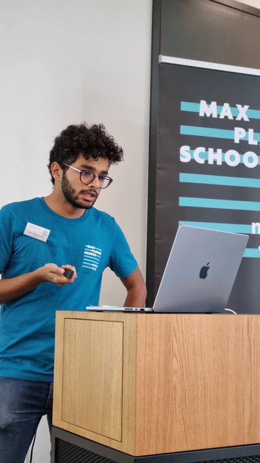 Tarun Mascarenhas stands at the lectern in an MtL T-shirt looking at the laptop with his slides, the laser pointer in his right hand.