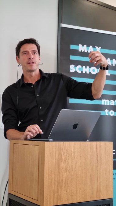 Close-up of the presentation by Hendrik Dietz. He is standing at the lectern in front of a roll-up banner of MPS MtL. He leans on the lectern with his right forearm, his left arm is raised and he underlines his explanations with a gesture.