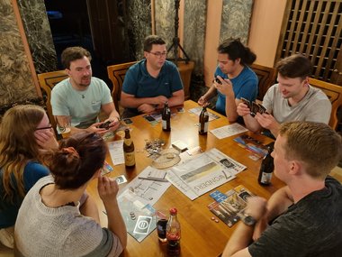 Five male and two female students are sitting around a wooden table playing a board game.