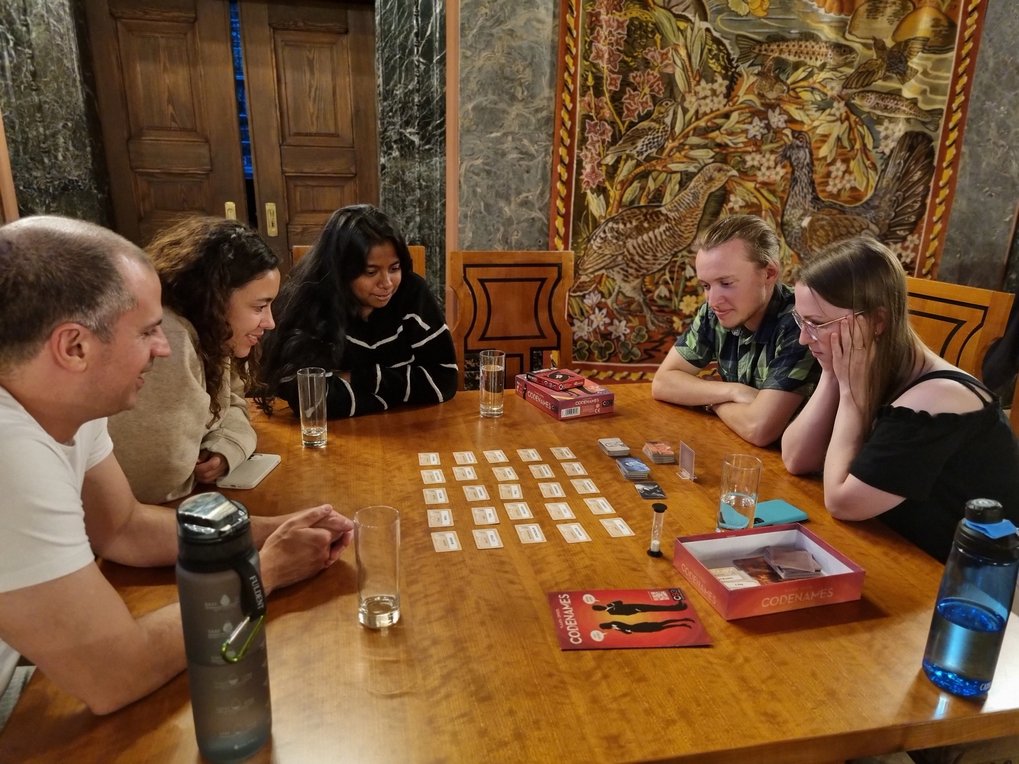 A group of five students, three women and two men, are sitting at a wooden table with a few rows of playing cards in the middle. They are looking intently at the playing cards, some of them smiling. Behind them is a tall, closed wooden door and a tapestry on a gray marble-look wall.