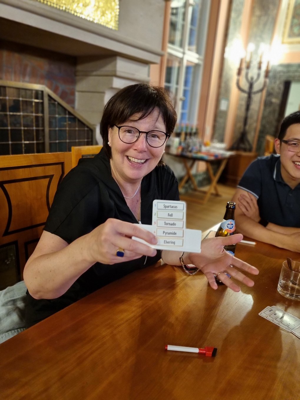 Professor Karin Jacobs sits at a wooden table and holds a small card with words from a board game up to the camera with a friendly grin. In the background you can see a chandelier.