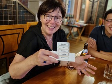 Professor Karin Jacobs sits at a wooden table and holds a small card with words from a board game up to the camera with a friendly grin. In the background you can see a chandelier.