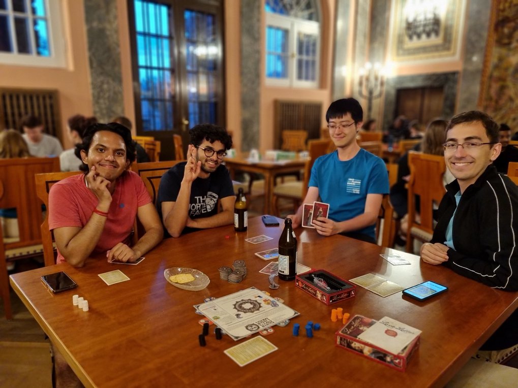 Four male students sit at a wooden table in a large, sumptuous hall and smile at the camera. One of them is holding playing cards and in front of them is a board game.