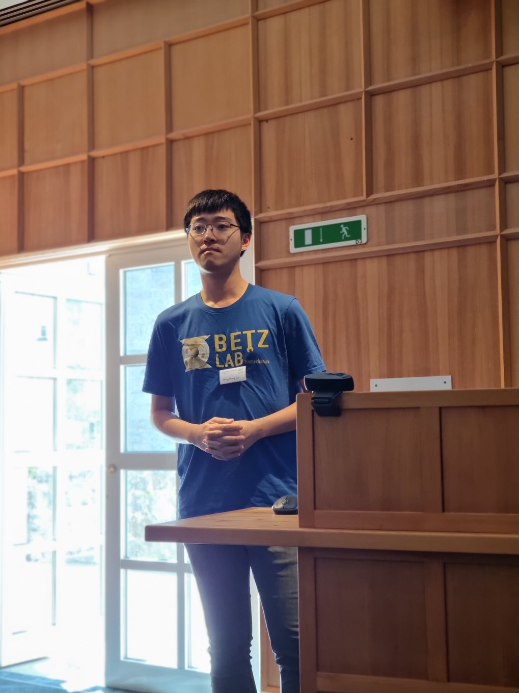 An MtL student stands with his hands clasped next to the lectern. He is wearing a shirt with the inscription "Betz Lab", the laboratory of faculty member Timo Betz.