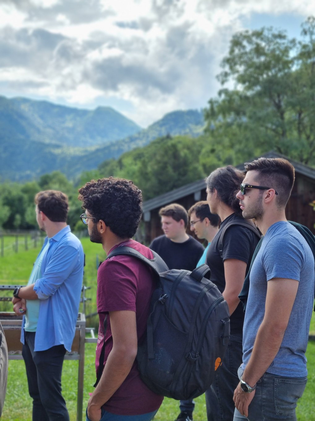 A group of students stand sideways to the camera in front of a green landscape. Mountains can be seen in the background, the sky is cloudy.
