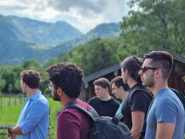A group of students stand sideways to the camera in front of a green landscape. Mountains can be seen in the background, the sky is cloudy.