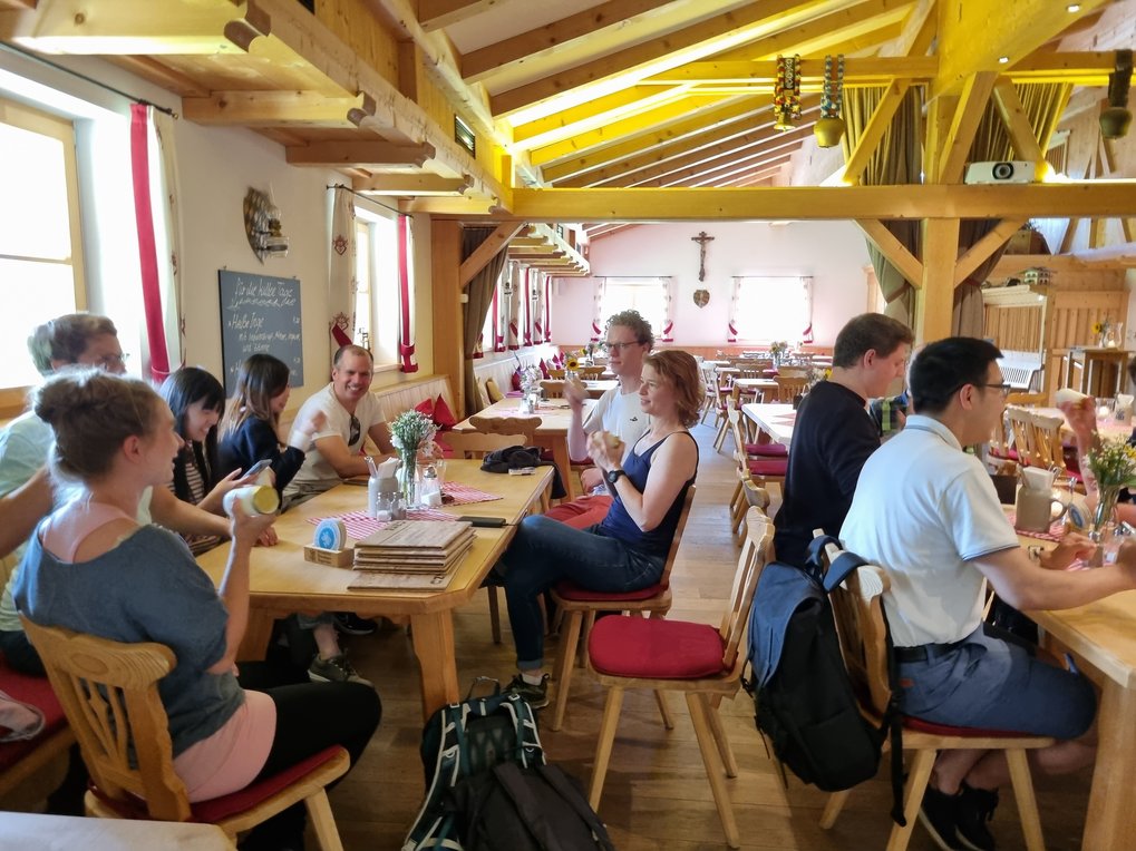 A group of participants sit at several wooden tables in a cozy, rustic dining room of a restaurant, which gives off a traditional alpine feel. The room has a warm atmosphere with wooden beams on the ceiling, walls painted in a light color, and small windows letting in natural light. The participants are shaking small glasses and thereby making butter.
