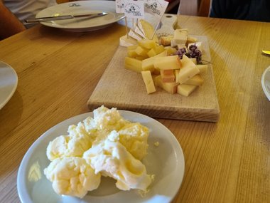 Homemade fresh butter on a round plate stands on a wooden table, behind it a wooden platter with a selection of sliced hard and soft cheeses.