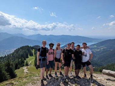 A group photo of six male and one female student. They are wearing hiking gear and backpacks and are standing on a mountain peak. Behind them is a green landscape with forests, mountains and a large lake in the distance. The sky is clear with few clouds.