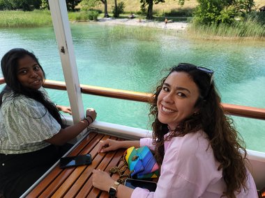 Zwei Studentinnen sitzen auf einem Boot, beide lächeln breit in die Kamera. Im Hintergrund sind das ruhige türkisfarbene Meer und ein grünes, von Bäumen gesäumtes Ufer zu sehen.