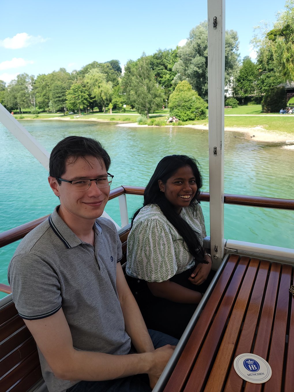 Zwei Studenten sitzen auf einer Holzbank auf dem Boot und lächeln in die Kamera. Hinter ihnen ist das ruhige, wunderschön türkisfarbene Meer zu sehen, sowie ein grüner Park mit Bäumen am Ufer.