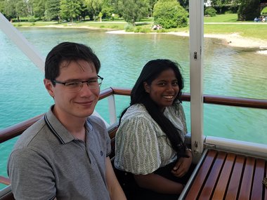 Zwei Studenten sitzen auf einer Holzbank auf dem Boot und lächeln in die Kamera. Hinter ihnen ist das ruhige, wunderschön türkisfarbene Meer zu sehen, sowie ein grüner Park mit Bäumen am Ufer.