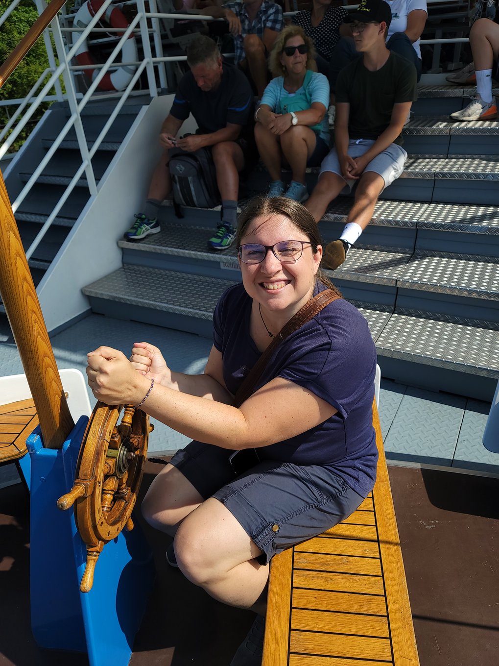 On a sunny day, a female MtL coordinator is smiling, slightly squinting, at the camera, holding onto a small, wooden ship's wheel as if she is steering the boat at the moment.