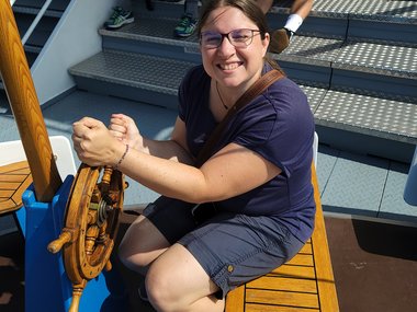 On a sunny day, a female MtL coordinator is smiling, slightly squinting, at the camera, holding onto a small, wooden ship's wheel as if she is steering the boat at the moment.