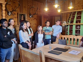A guided tour of the castle offered insights into its history and tragic fate of creator Friedrich Attenhuber A diverse group of students stand in a rustic wooden room adorned with antler trophies and a portrait painting. They are attentively listening to the castle manager giving a tour.