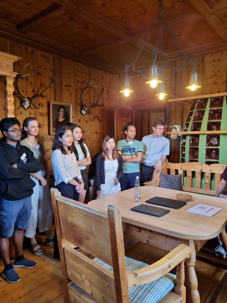 A diverse group of students stand in a rustic wooden room adorned with antler trophies and a portrait painting. They are attentively listening to the castle manager giving a tour.