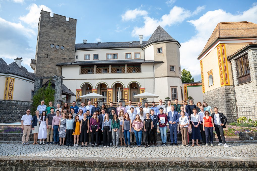 Group picture of the around 50 attendees in the courtyard with cobblestone paving. In the back, you can see Ringberg Castle: An elegant historical building with a mix of medieval and modern architectural elements. The castle features a prominent stone tower, arched windows, and decorative facades with yellow and red accents. The participants are a diverse mix of men and women, most of them dressed in business casual attire. The weather is sunny, but some clouds can be seen in the sky.