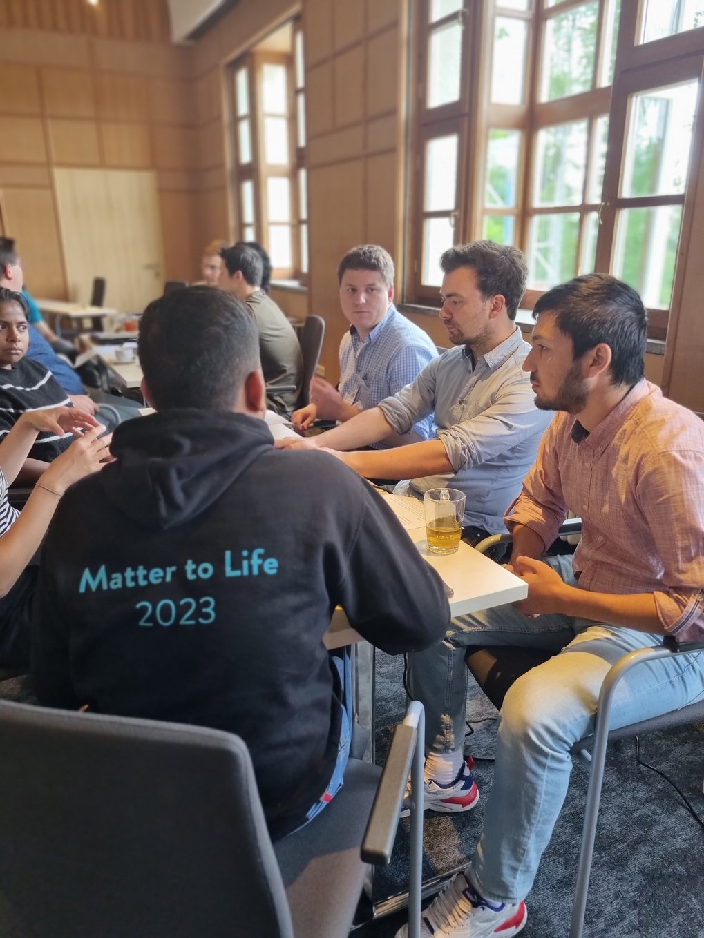 A group of male students are engaged in a discussion around a table in a room with wooden interiors. One person's back is visible with a black hoodie that reads 'Matter to Life 2023'. The facial expressions show attentiveness and focus.