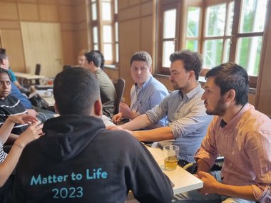 A group of male students are engaged in a discussion around a table in a room with wooden interiors. One person's back is visible with a black hoodie that reads 'Matter to Life 2023'. The facial expressions show attentiveness and focus.