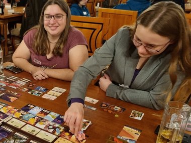 Two female participants sit at a wooden table and play a board game