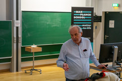 Matter to Life scholarship holder Jörg Stülke stands in the Physics lecture hall of the University of Göttingen and talks about his research. Behind him is a large green board and a Matter to Life roll up.