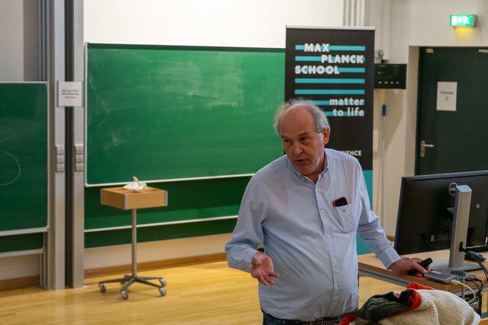 Matter to Life scholarship holder Jörg Stülke stands in the Physics lecture hall of the University of Göttingen and talks about his research. Behind him is a large green board and a Matter to Life roll up. 