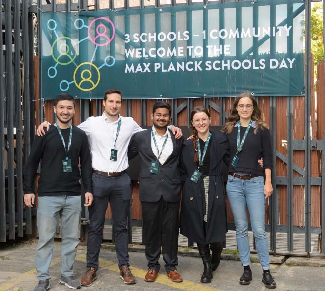 Three Matter to Life PhD candidates and two female PhD candidates arm in arm in front of a banner for the Max Planck School Day 2023