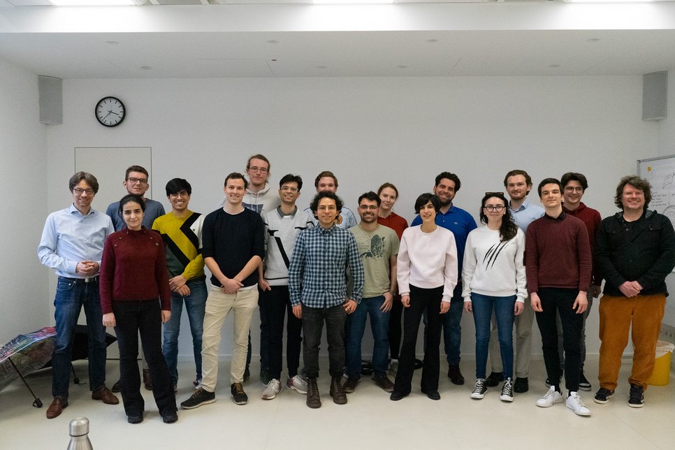 Sixteen students and two educators from the Max Planck School Matter to Life smile into the camera. They are standing in front of a white wall on which a clock can be seen