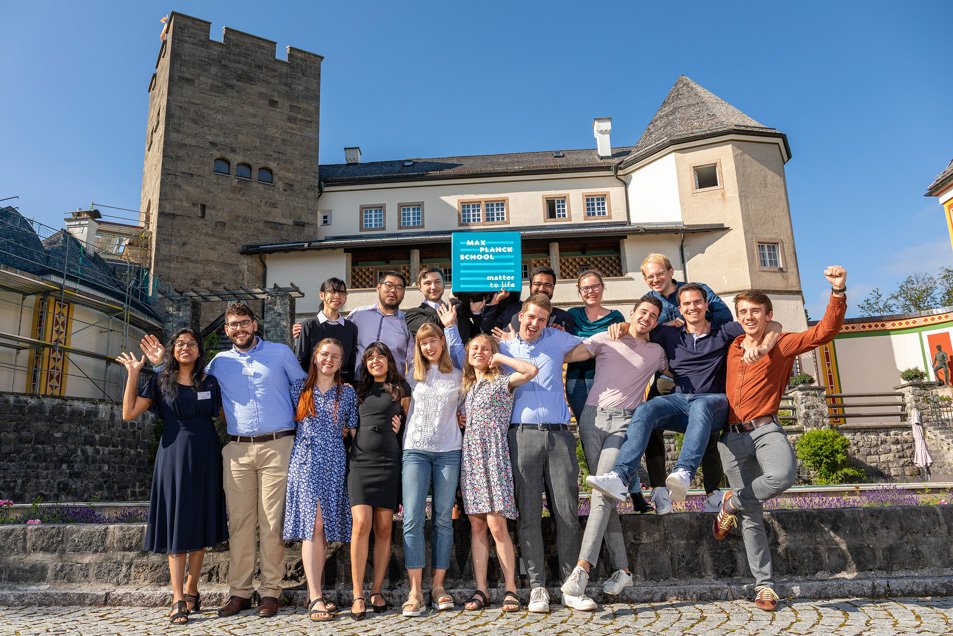A group of students pose happily for a group photo, with two students holding up a seating cube with the Matter to Life logo. Ringberg Castle can be seen in the background. 