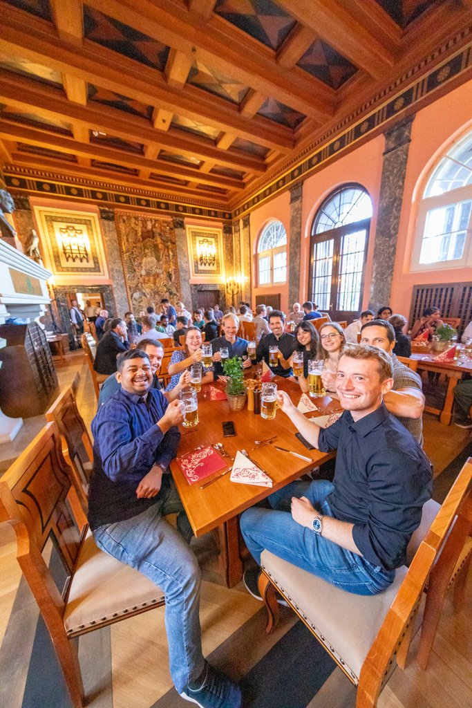A group of nine students sit together at a table in the dining hall at Ringberg Castle, smiling and raising their glasses of beer to the camera. 