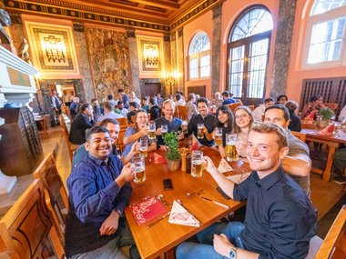 A group of nine students sit together at a table in the dining hall at Ringberg Castle, smiling and raising their glasses of beer to the camera.