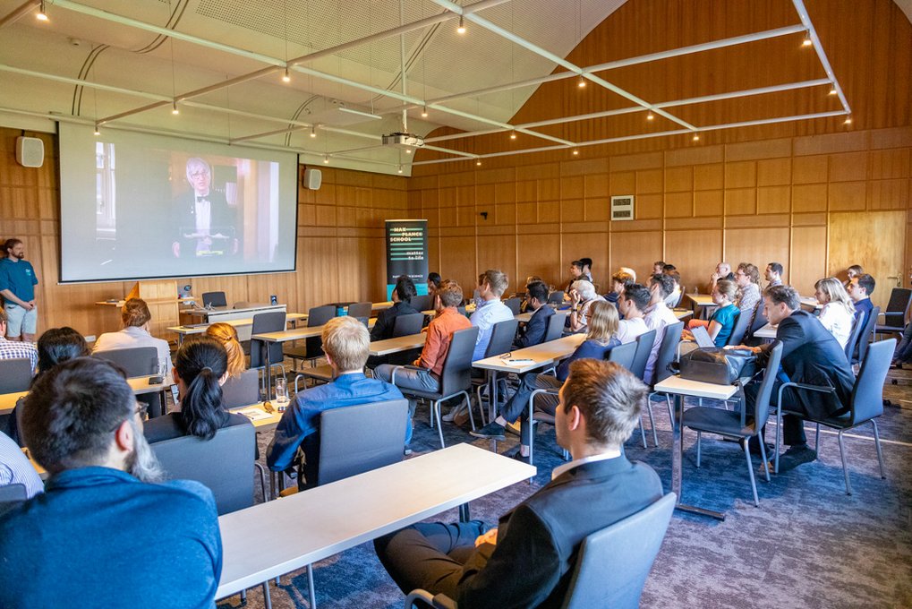 PhD candidates and professors sit in a lecture room and look at a screen showing Professor Tolan, President of the University of Göttingen. 